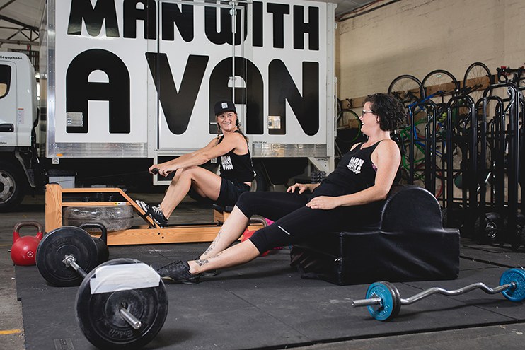 Two women in Man With A Van uniform work out on the company gym equipment.