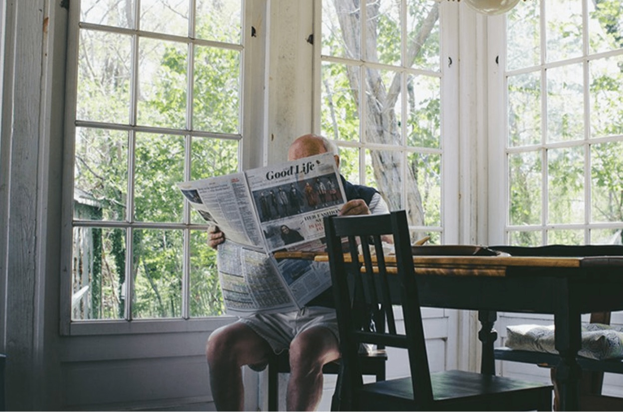 An elderly man, sitting at a dining table, reading a newspaper.